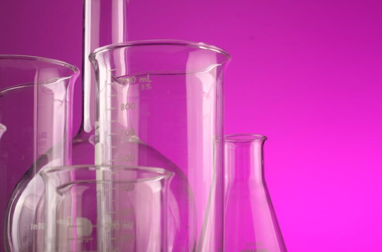 Several clear glass laboratory beakers and flasks, some with measurement markings, are grouped together against a bright pink background.