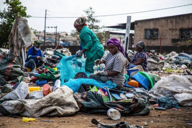 Several people sort through piles of plastic waste and recyclables in an outdoor area filled with assorted garbage, with buildings and fencing visible in the background.