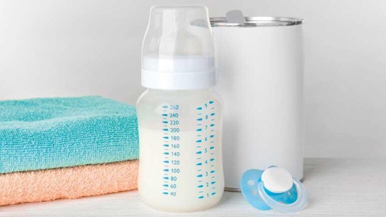 A baby bottle filled with milk stands in front of a white canister, beside two folded towels in blue and peach, and two blue pacifiers on a white surface.