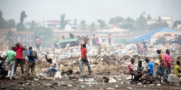 People are collecting and sorting through piles of waste in a large open landfill, with buildings and trees visible in the background under a hazy sky.