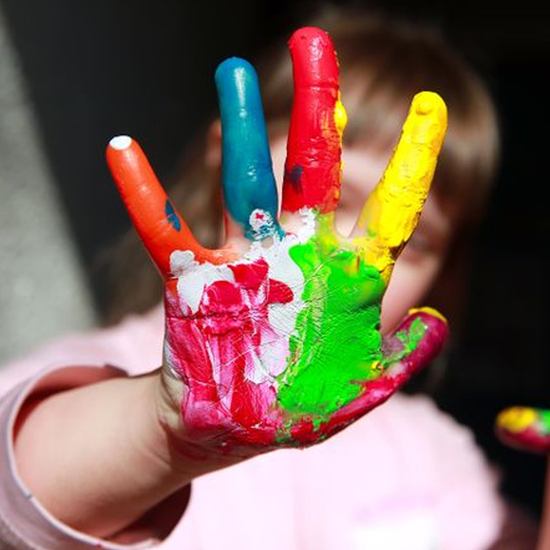 A child holds up a hand covered in colorful paint, with each finger painted a different color and the palm showing a mix of red, white, and green paint. The childs face is blurred in the background.