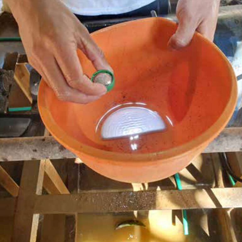 A person holds a small round object over an orange bowl filled with water. The bowl is placed on a wooden structure, and reflections are visible on the water’s surface.
