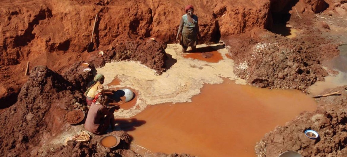 Three people are standing and crouching in a muddy, reddish-brown open pit, possibly mining or panning, with bowls and plates around them and pools of murky water filling the pit.