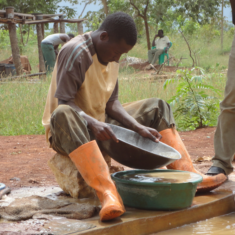 A man sitting on a rock wears orange boots and washes a pan in a green tub of muddy water outdoors, with trees and other people in the background.