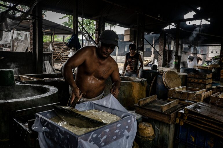 A shirtless man wearing a cap uses a large metal scoop to transfer a wet, white substance into a rectangular container inside a dimly lit, industrial setting. Another person works in the background amid various tools and materials.