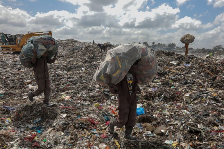 Two people carry large bags filled with collected items on their backs while walking through a landfill covered in trash and debris under a cloudy sky; a bulldozer operates in the background.