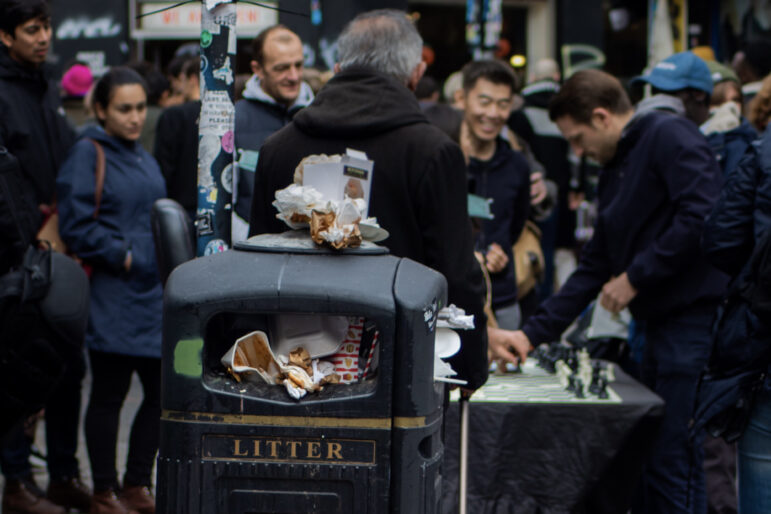An overflowing litter bin with food wrappers and boxes in focus. In the background, people are gathered around a street chess game on a crowded city sidewalk.