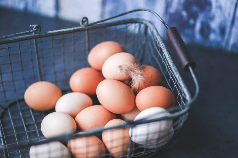 A wire basket containing several brown and white eggs. A small feather is resting on top of some of the eggs, and the background appears to be a rustic, textured surface.