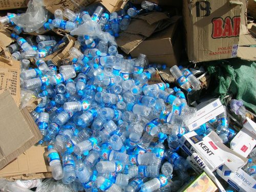 A large pile of empty plastic water bottles, flattened cardboard boxes, and scattered packaging materials, suggesting waste or recycling. The bottles are mostly clear with blue caps and labels.