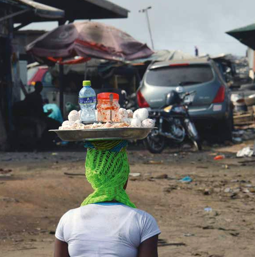 A person with a green net scarf carries a tray of bottles, jars, and white items on their head in a busy outdoor market with cars, motorcycles, and tents in the background.