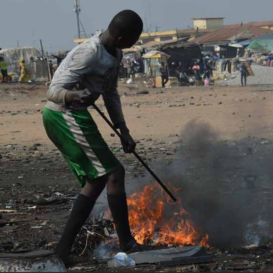 A person wearing a gray shirt, green shorts, and black boots uses a metal rod to tend to a small fire with black smoke outdoors in a cluttered area, with buildings and people in the background.