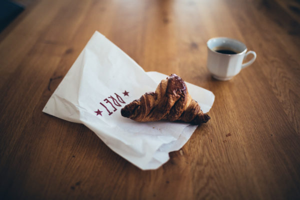 A chocolate croissant sits on a crumpled Pret-branded paper bag on a wooden table, next to a white cup filled with black coffee.