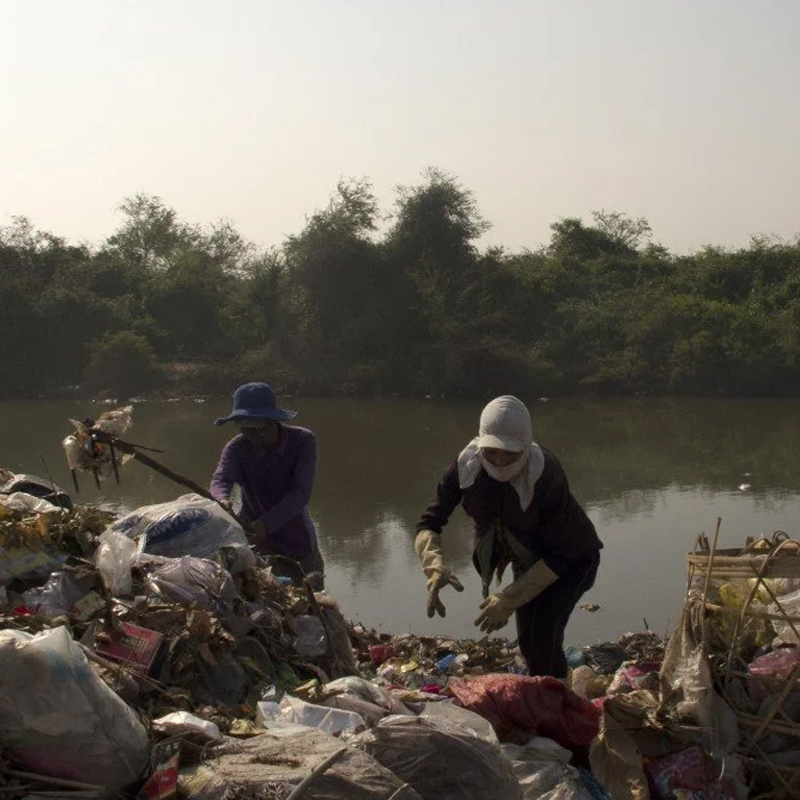 Two people wearing hats and gloves are sorting through a large pile of garbage near a body of water, with trees and bushes in the background under a clear sky.