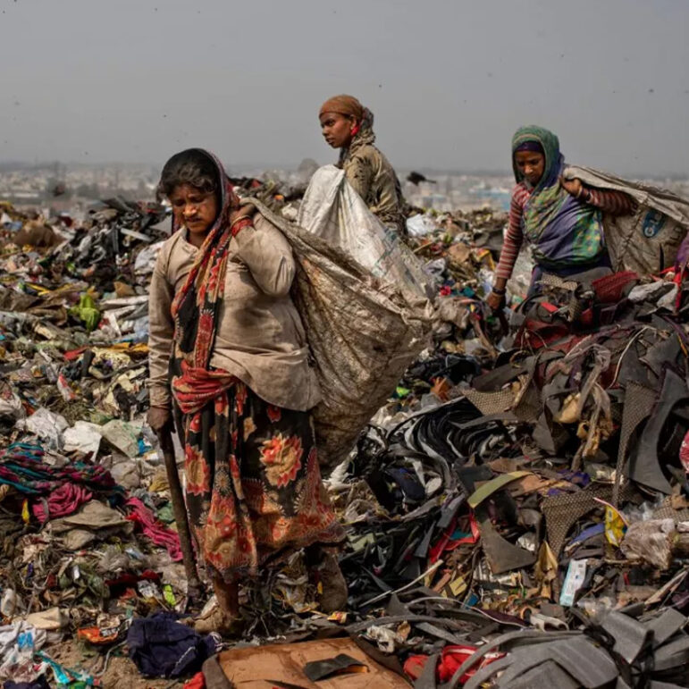 Three women walk through a large pile of garbage, carrying sacks on their backs and wearing worn clothing. The environment is filled with discarded materials under an overcast sky.