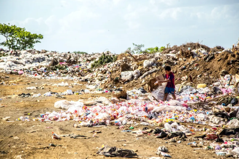 A person stands among piles of plastic waste and garbage at a large landfill site under a partly cloudy sky, with scattered trash covering the ground.