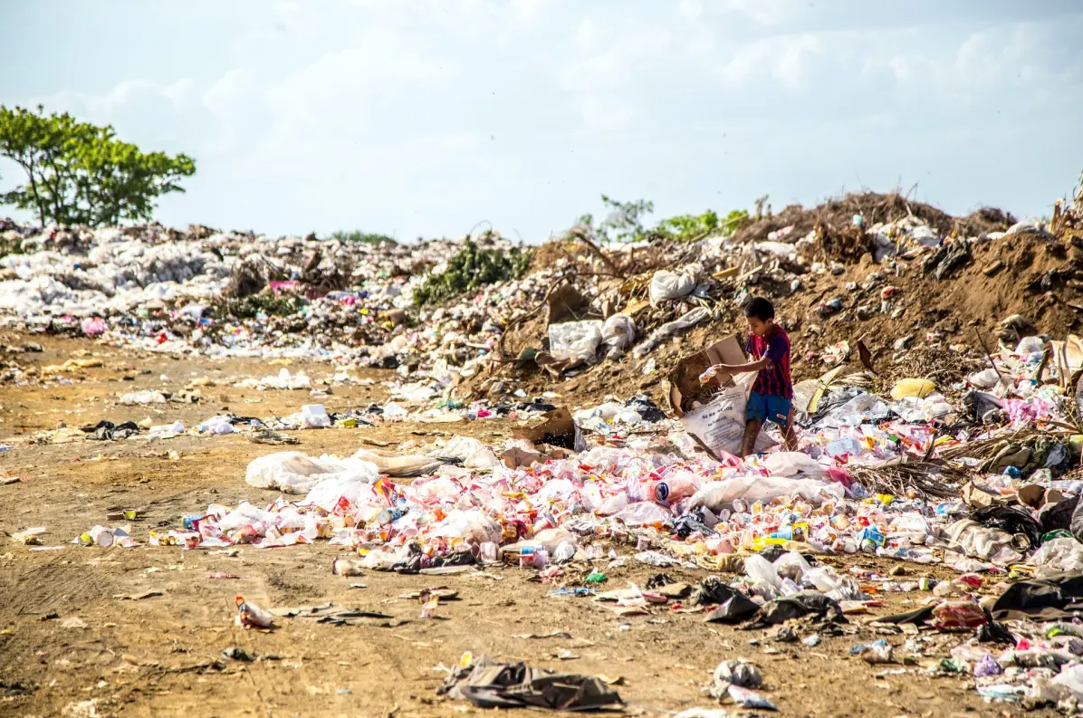 A person stands among piles of plastic waste and garbage at a large landfill site under a partly cloudy sky, with scattered trash covering the ground.