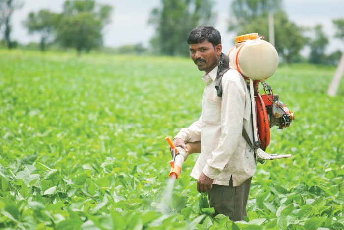 A man stands in a green field wearing a white shirt and carrying a pesticide sprayer on his back, spraying crops with a hose. Trees and plants fill the background under a cloudy sky.