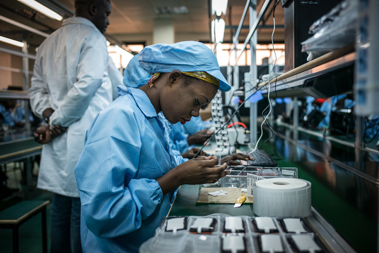 A woman in a blue lab coat and hair cover assembles electronic components at a workstation in a factory. Other workers in similar attire are seen working in the background.