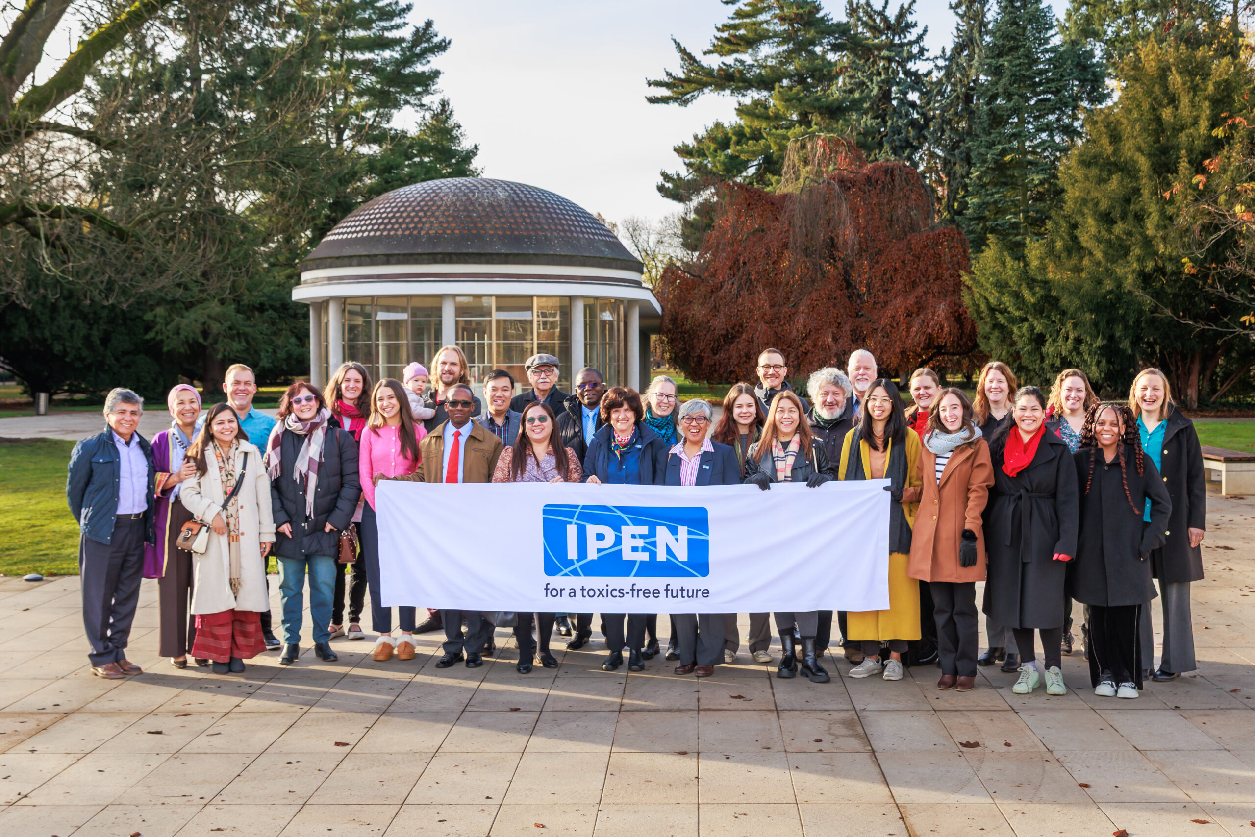 A group of about 30 people stands outdoors in front of a small pavilion, holding a large banner that reads IPEN for a toxics-free future. The group is smiling and dressed in winter clothing. Trees are visible in the background.