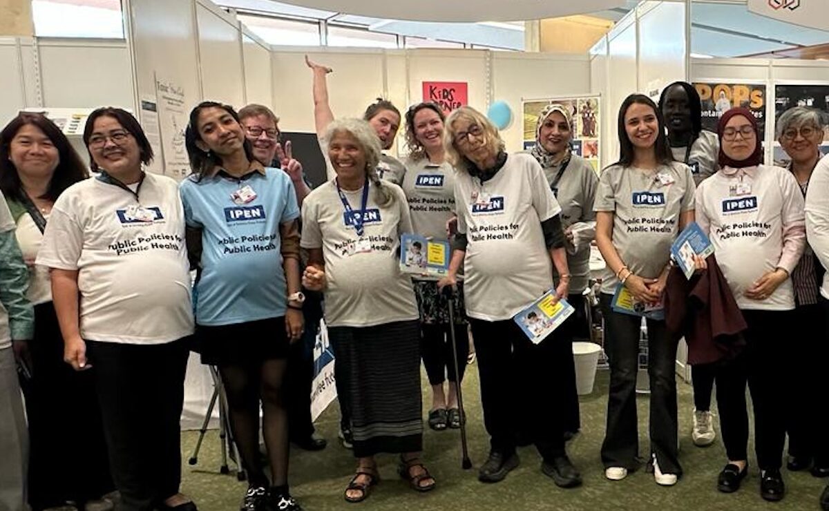 A group of people stands and smiles in front of a booth labeled “International Pollutants Elimination Network.” Several are wearing matching shirts and holding pamphlets. The booth is set up in an indoor event space.