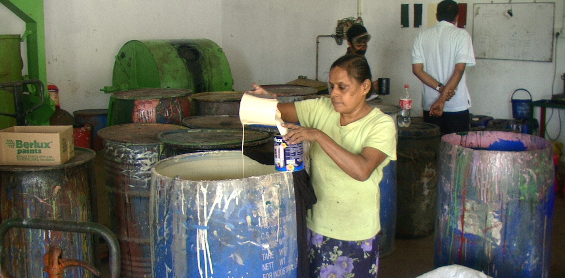 A woman pours white paint from a jug into a smaller can in a room filled with large paint barrels. Other people work in the background near equipment and a wall with various objects.