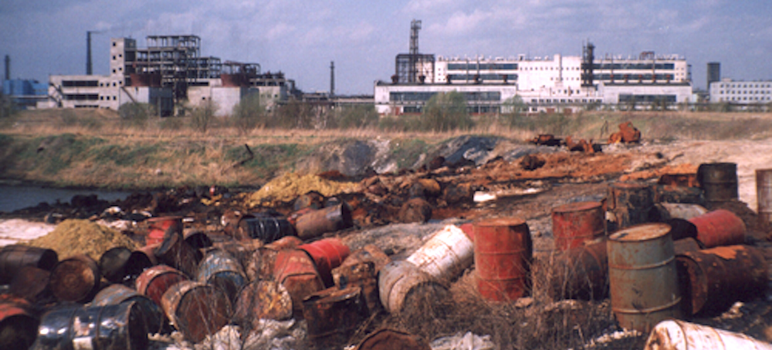 Several rusted and damaged barrels are scattered on the ground near a body of water, with an industrial facility and some vegetation visible in the background under a partly cloudy sky.