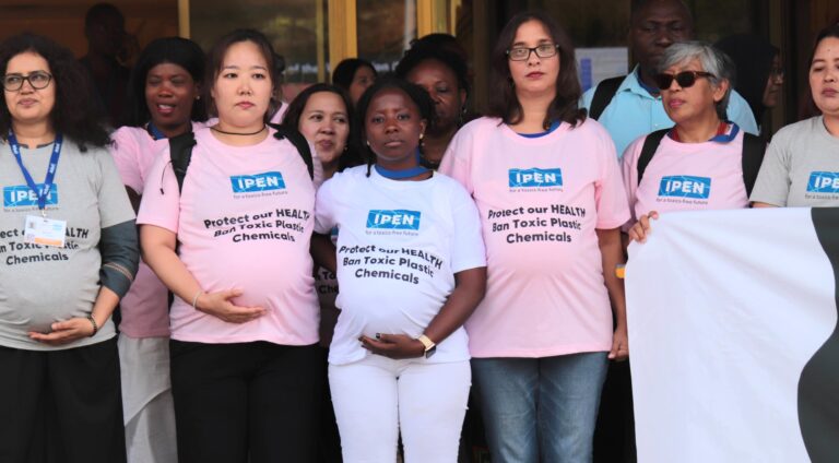 A group of people, some appearing to be pregnant, stand together wearing shirts that read Protect our health, ban toxic plastics, chemicals. They are gathered in front of a building, participating in a demonstration.