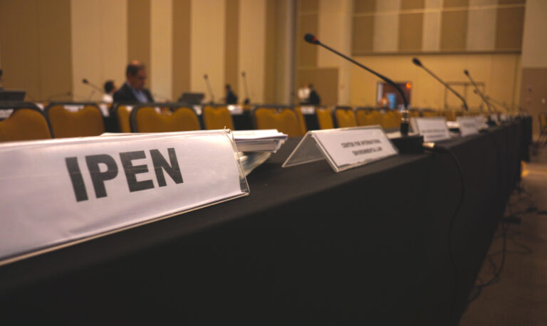 A row of tables with nameplates and microphones in a conference room. The nameplate in the foreground reads IPEN. Several empty chairs and a few people sitting in the background are visible.