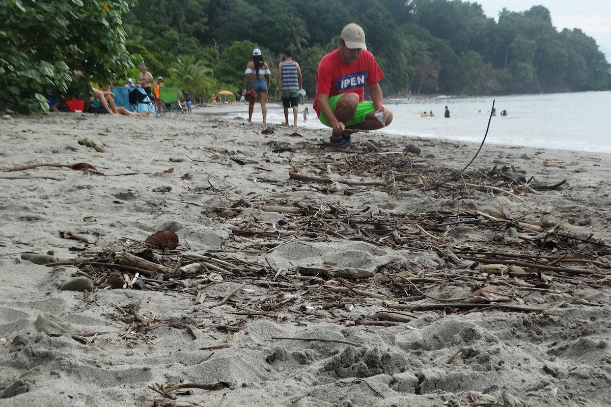 A person in a red shirt and beige cap is squatting on a sandy beach near driftwood and debris. Other people are walking and swimming in the background, with trees lining the shore.