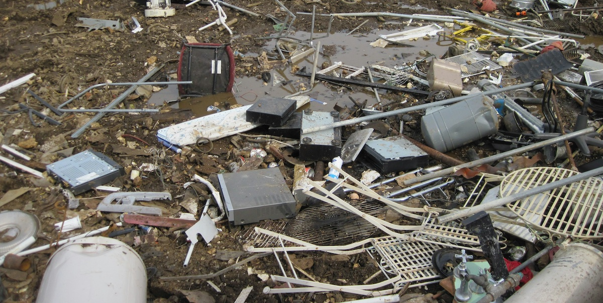 A muddy area is scattered with electronic waste, metal parts, wires, plastic containers, and household debris, creating a cluttered and chaotic junkyard scene.