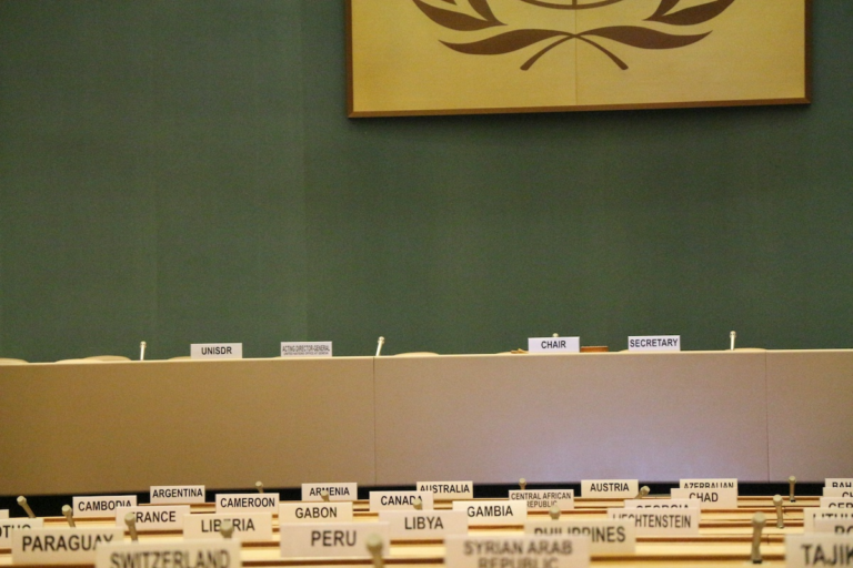 A conference room with empty chairs behind a desk labeled UNISDR, CHAIR, and SECRETARY. In the foreground are nameplates for various countries, including Argentina, France, and Paraguay. A United Nations emblem hangs above.