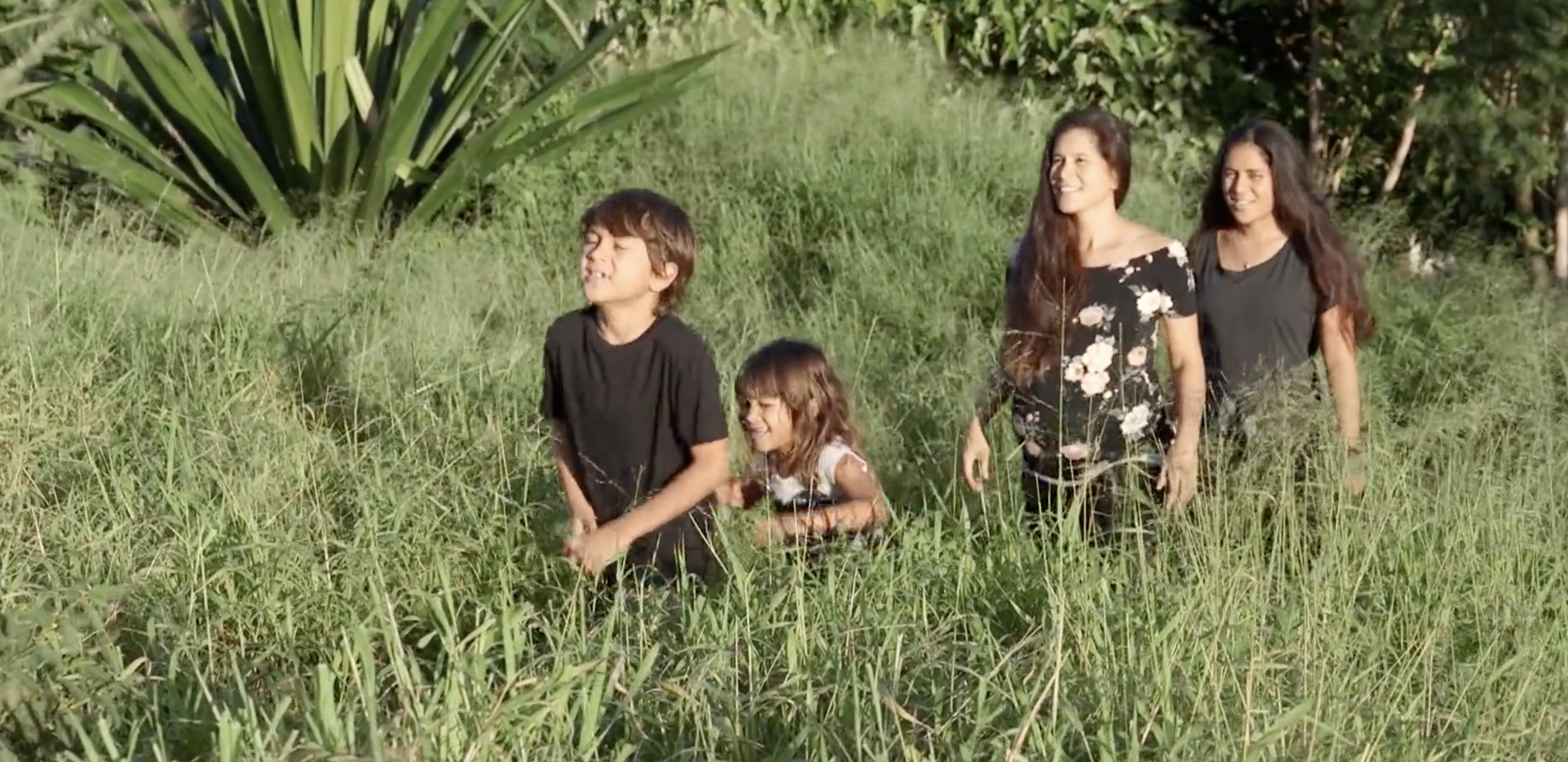 A pregnant mom with her children walking together in tall green grass, looking ahead and smiling. The sunlight is bright, and surrounding plants create a natural, outdoor setting.