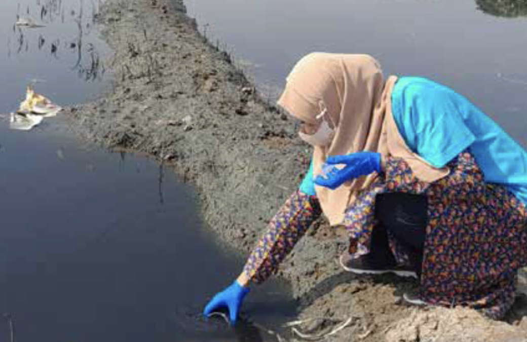 A person wearing a hijab, face mask, and blue gloves crouches by the edge of a polluted body of water, collecting a sample with a small container.