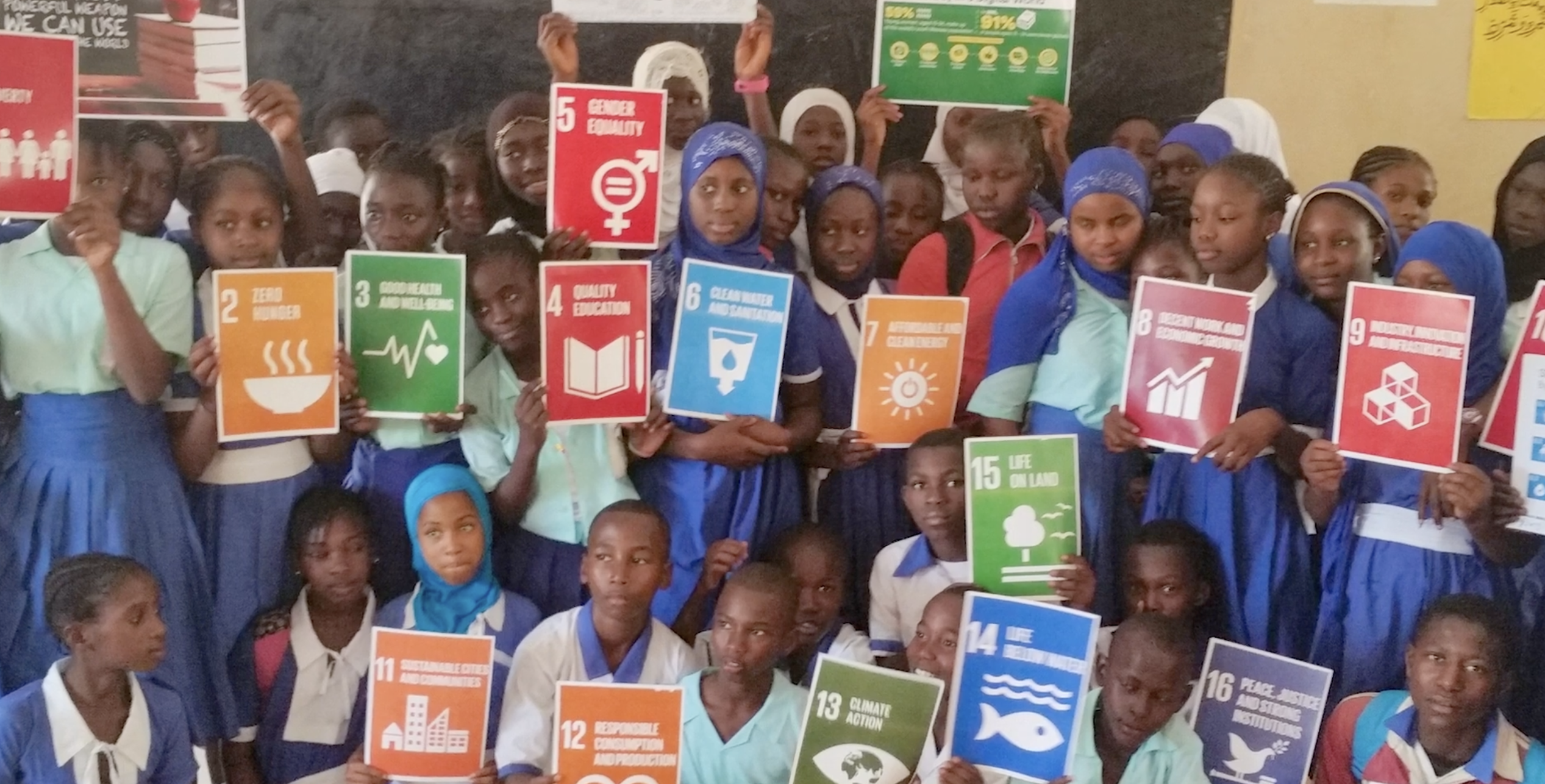 A group of schoolchildren in uniform hold up colorful signs displaying various United Nations Sustainable Development Goals inside a classroom.