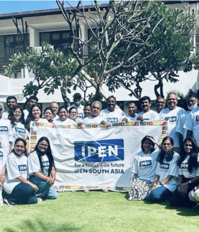 A group of people in matching IPEN t-shirts pose outdoors, holding a large banner that reads “IPEN for a toxics-free future IPEN South Asia,” with a building and trees in the background.