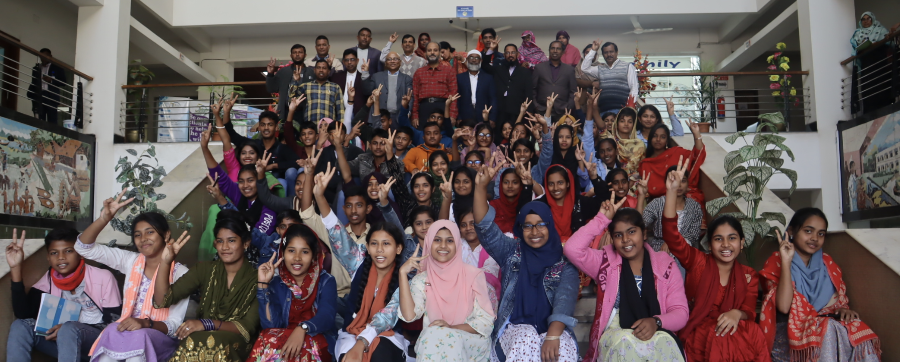 A large group of people, including adults and children, pose together on indoor stairs. Many are smiling and making peace signs with their hands. The setting appears to be a public or community building.