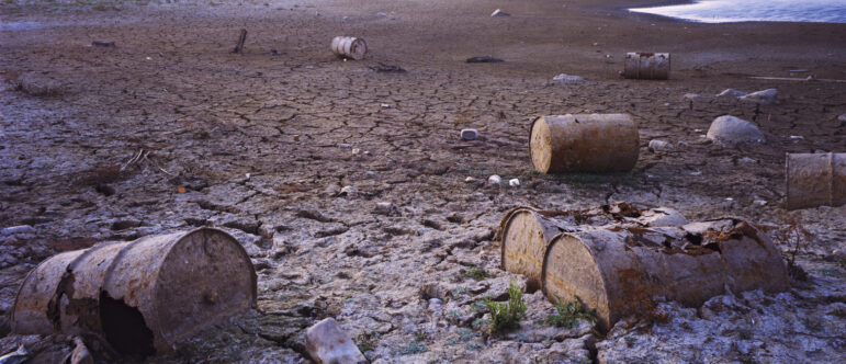 Rusty metal barrels lie scattered on cracked, dry earth near the edge of a receding body of water, with distant mountains visible under a hazy sky. The landscape appears barren and desolate.