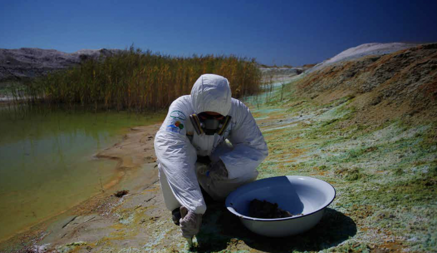 person in a biohazard suit and gas mask taking samples in a field