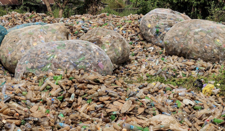 Large piles of discarded plastic bottles cover the ground, with several transparent bags filled with more plastic bottles among them. There is sparse greenery visible around the waste.