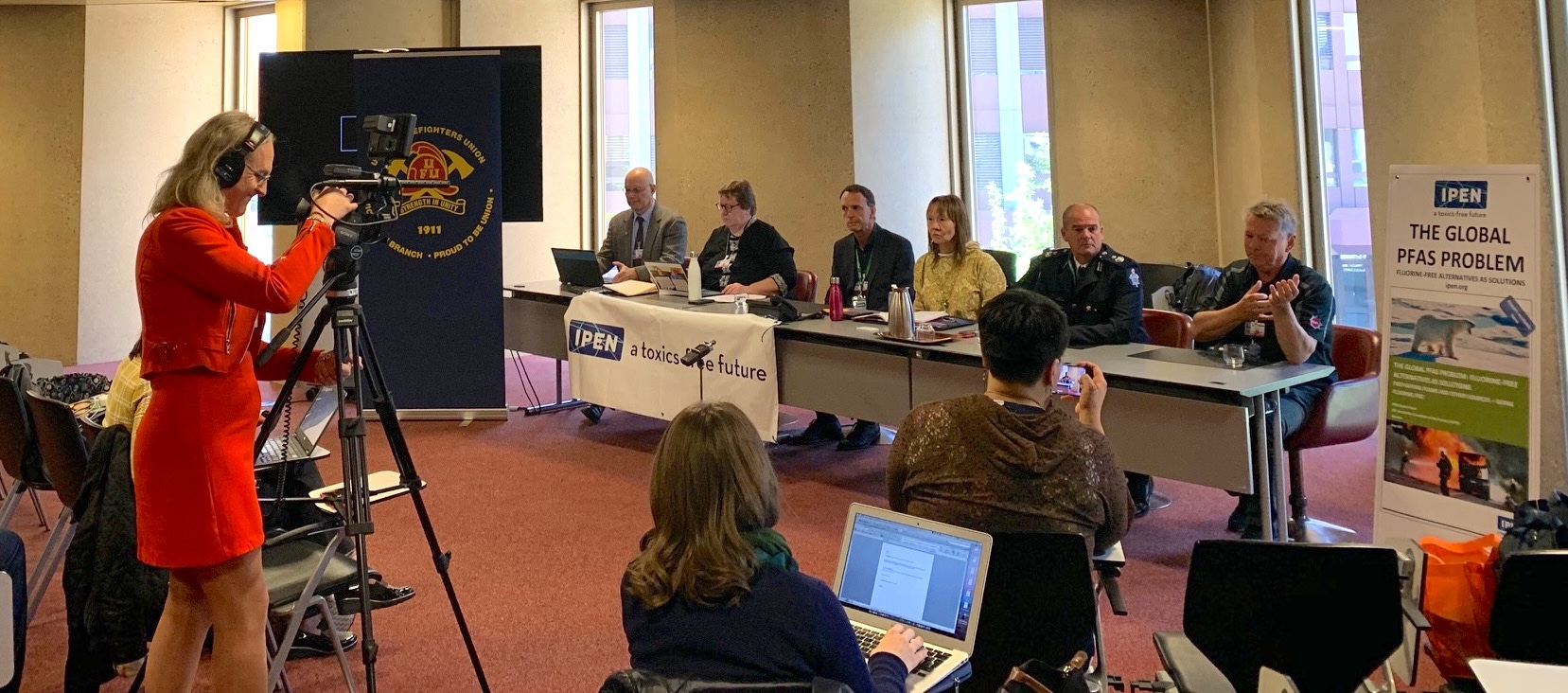 A woman in a red dress films a panel of six people seated at a long table during a conference or discussion. Audience members are seated nearby, some using laptops. A poster about PFAS is displayed on the right.
