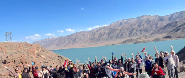 A large group of people pose with arms raised near a turquoise lake, rocky terrain, and mountains under a clear blue sky.