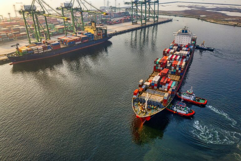A large cargo ship loaded with shipping containers is guided by tugboats near a port with cranes and stacked containers visible onshore. The scene is set during daylight.