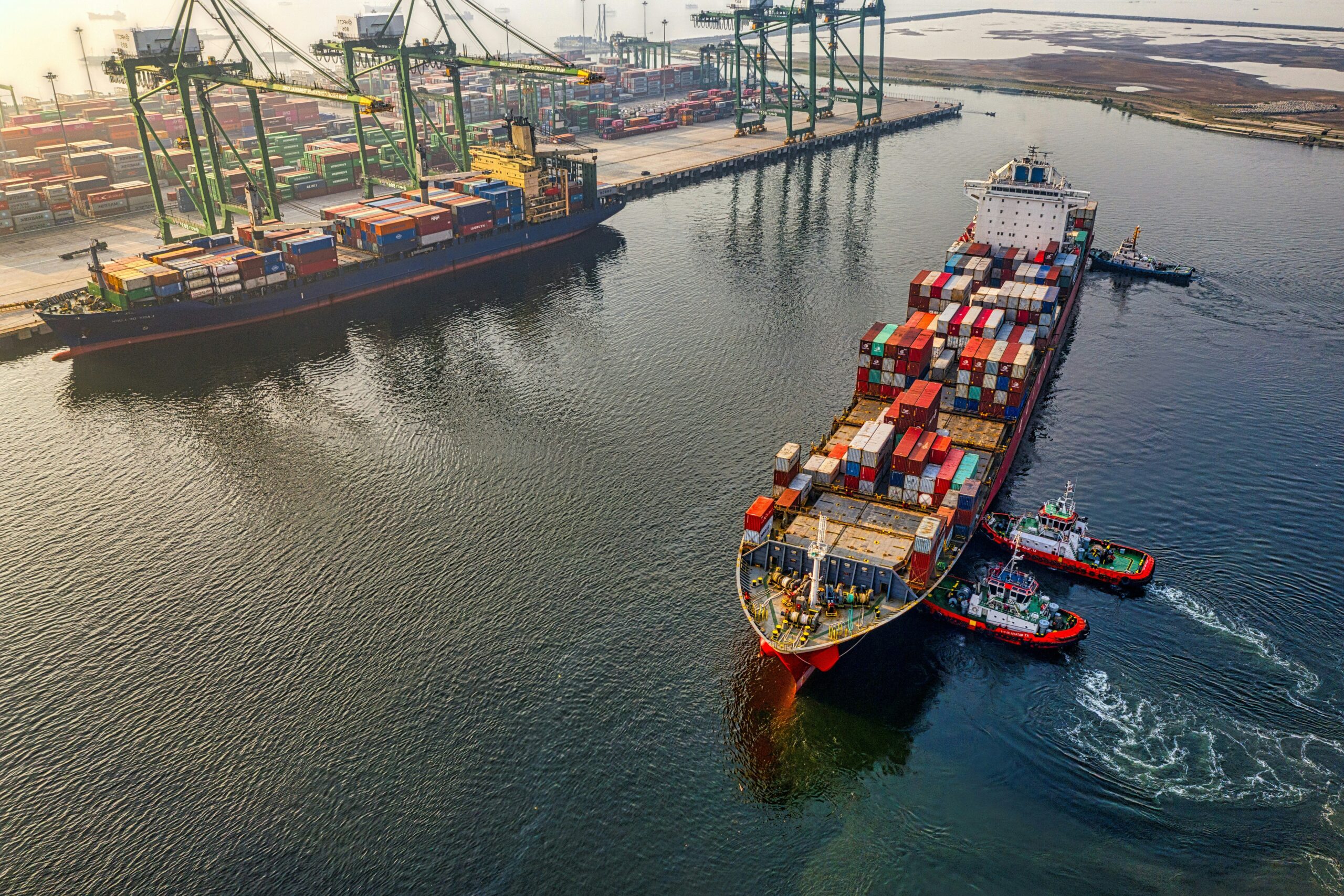 A large cargo ship loaded with shipping containers is guided by tugboats near a port with cranes and stacked containers visible onshore. The scene is set during daylight.