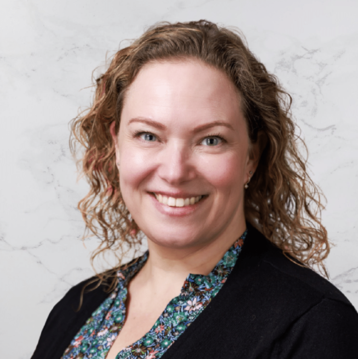 A woman with curly blonde hair smiles at the camera. She is wearing a dark cardigan over a floral-patterned blouse and is posed in front of a light-colored, marbled background.