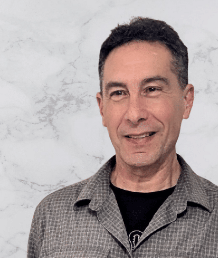 A middle-aged man with short dark hair, wearing a gray plaid shirt over a black T-shirt, stands in front of a light gray marble-patterned background, looking slightly to his left and smiling.