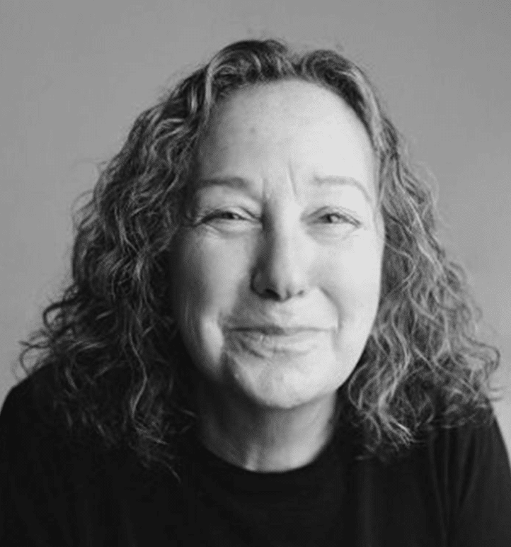 A woman with curly hair smiles gently at the camera. She is wearing a dark top and the background is plain and neutral. The image is in black and white.
