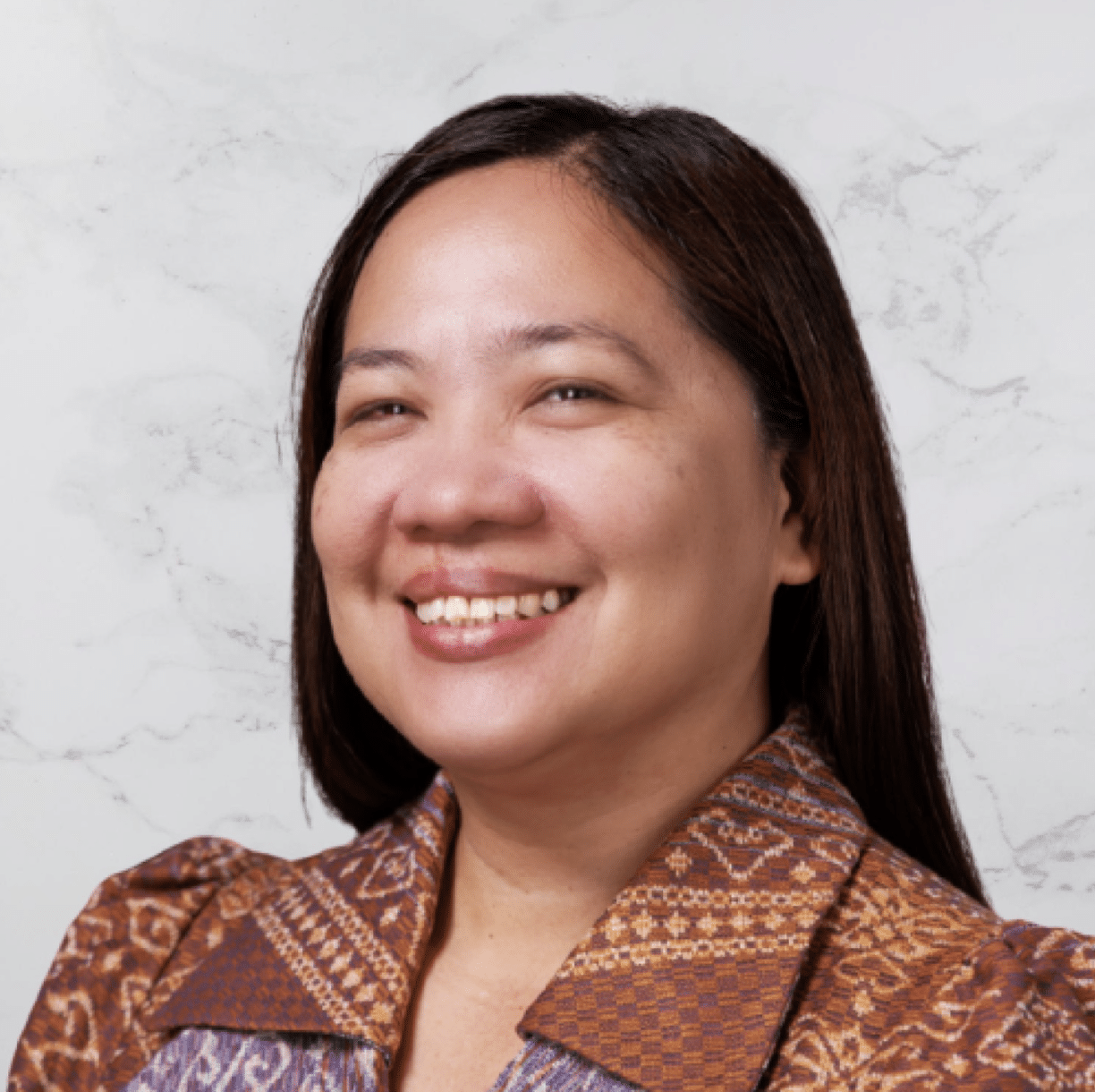 A woman with straight, dark brown hair is smiling. She is wearing a patterned brown blouse and is posed against a light-colored, marbled background.