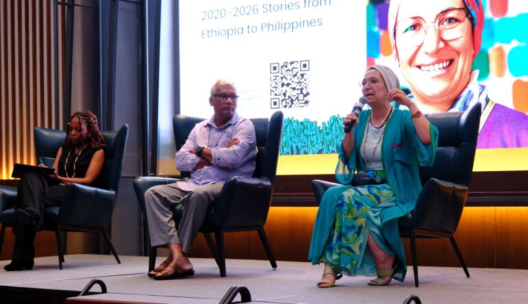 Three people are seated on a stage panel. A woman in a blue and green outfit is speaking into a microphone. Behind them is a screen with a large photo of a smiling woman, text, and a QR code.