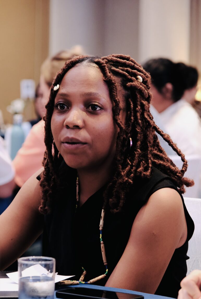 A woman with medium brown skin and long, twisted hair sits at a table, facing forward. She is wearing a sleeveless black top and a beaded necklace. Several people are visible in the blurred background.