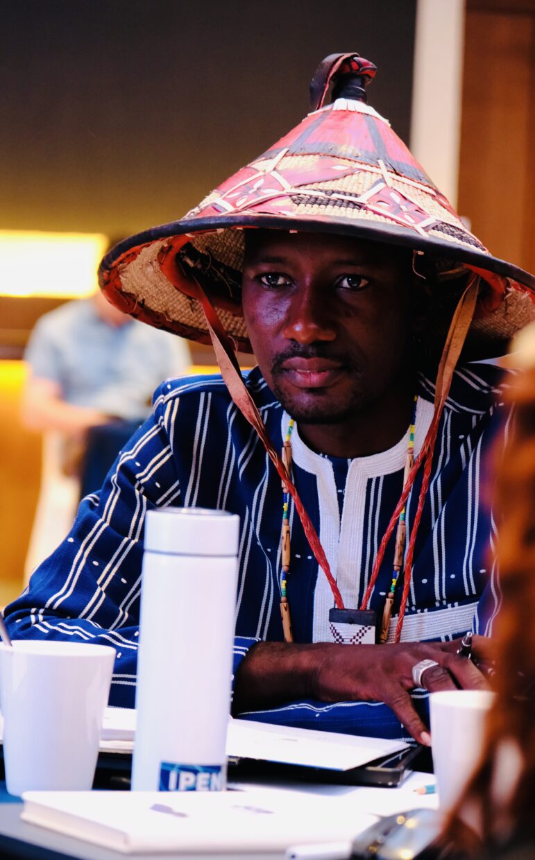 A man wearing a traditional woven hat and a striped shirt sits at a table with a white thermos, cups, and papers in front of him, looking attentive. The background is softly blurred.